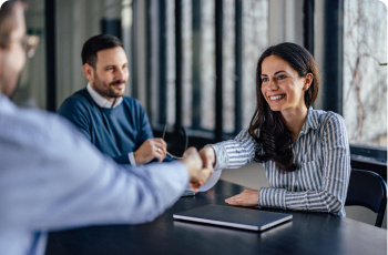 Three people sat at a table in an office, two people are shaking hands.