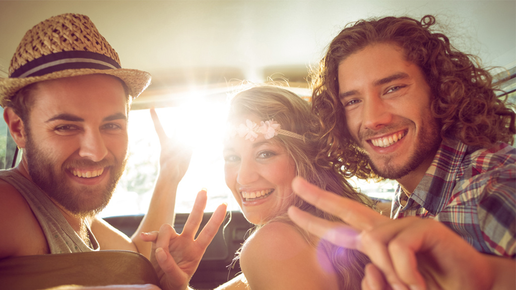 Three friends in a car smiling and wearing summer clothing, looking back at the camera the sun shining behind them.