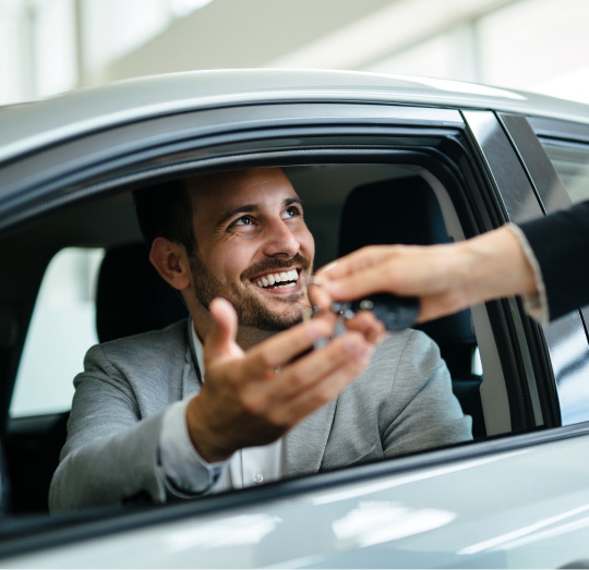 A man in a grey suit sitting in a car with the window open, a person is handing him some keys.