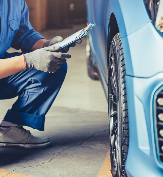 A person squatted down by a car wheel, the person is wearing a pair of plastic gloves and holding a clipboard.
