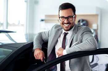 A man in a grey suit standing by a car with the car door open.