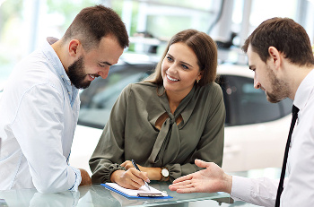 Two men and a woman talking in a car showroom, the woman is taking notes.