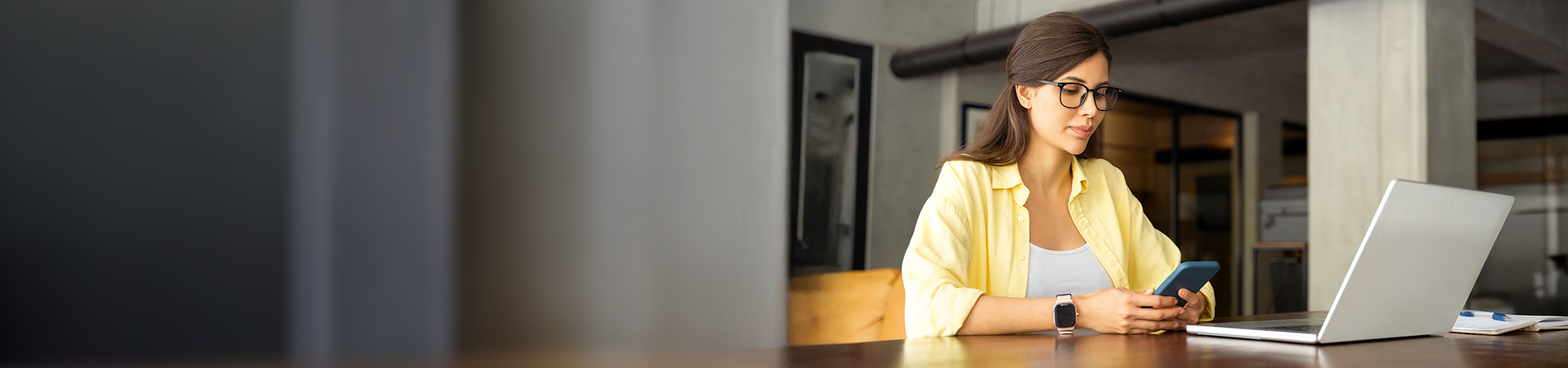 A woman sat at a desk on her mobile phone with a  laptop in front of her.
