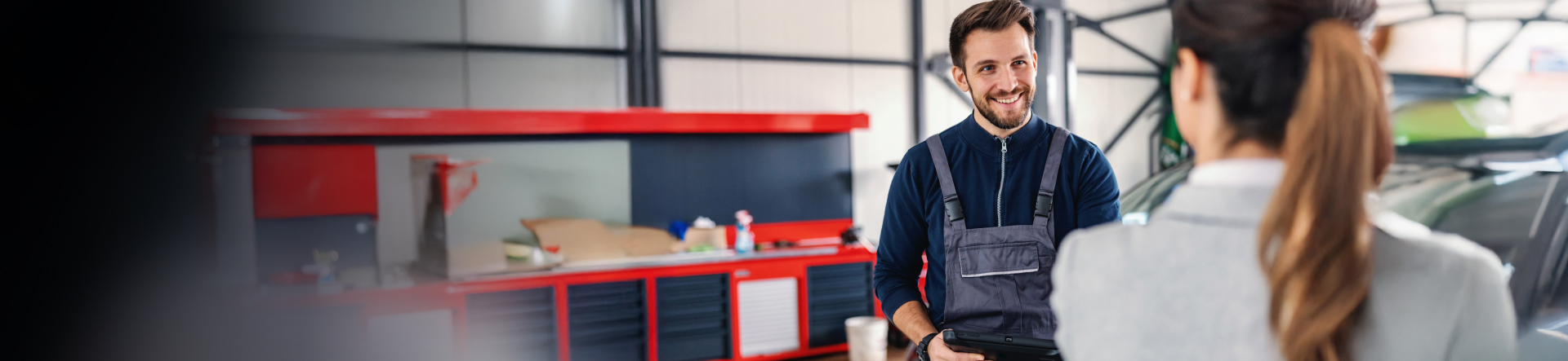 A man in overalls at a garage speaking to a woman with a ponytail wearing a pale grey suit.