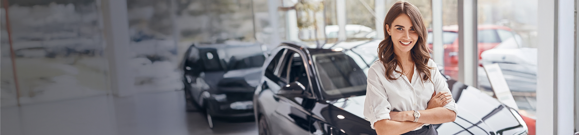 A woman stood in a car forecourt, wearing office clothing smiling and folding her arms. 