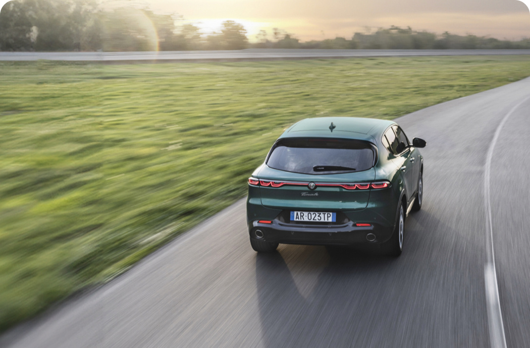 A car driving down a road with green grass to the side and trees on the skyline in front of it.