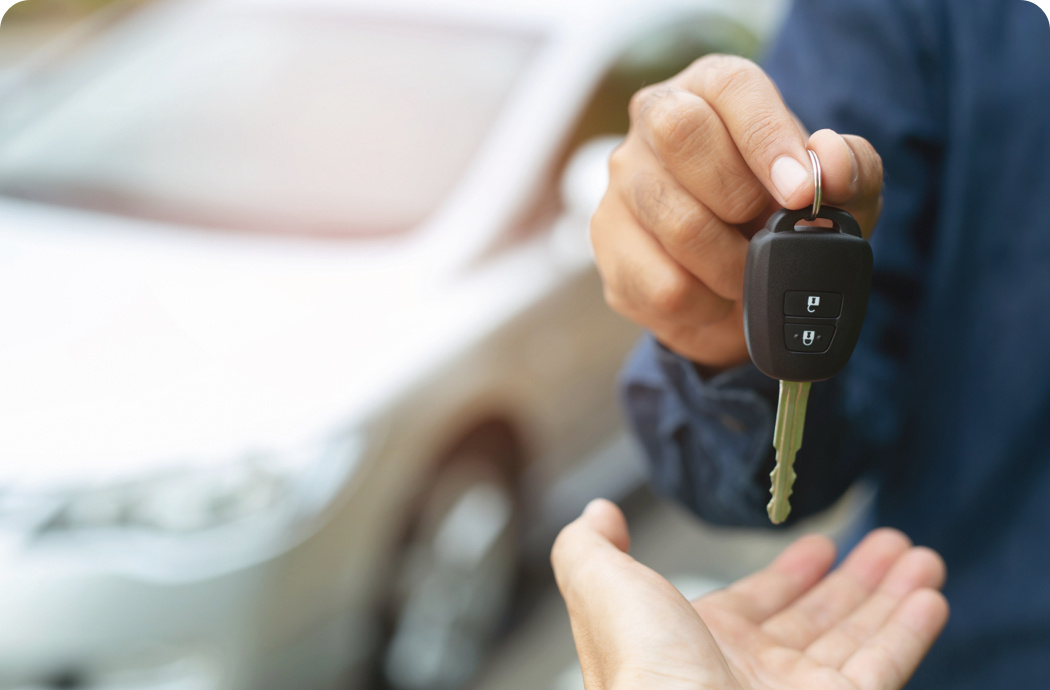 A person wearing overalls handing over a car key to someone. There is a car in the background.