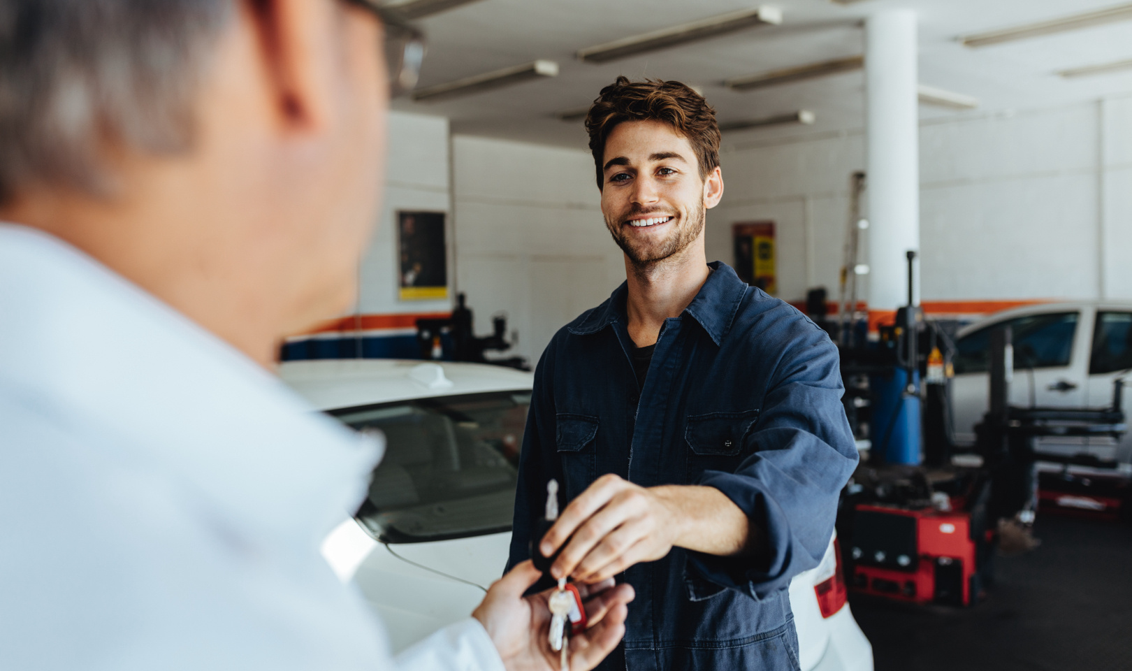 A man in overalls in a garage passing another man a set of car keys.