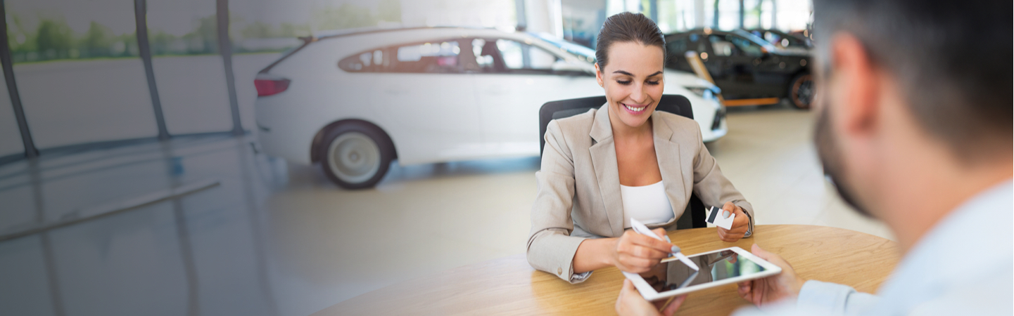 A woman signing something on an ipad whilst holding a bank card. There is a man sat across the desk from her and a car in the background. 