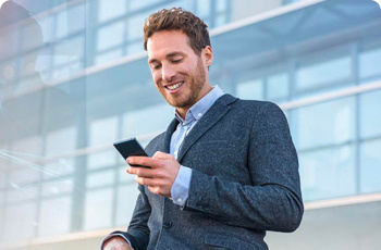 A man standing outside an office block looking at this mobile phone and smiling.