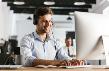 A man in an office environment with a headset on looking at a screen and smiling.