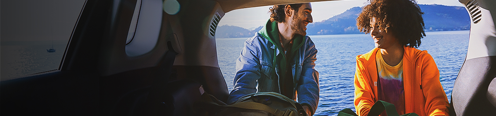 A man and a woman with waterproofs on getting some things out of a boot of a car. The sea and some mountains are in the distance.