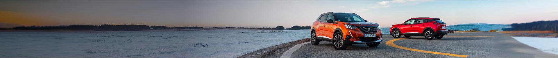 Two cars parked on tarmac, the sea is in the background.